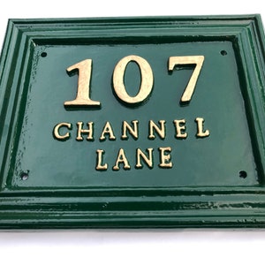 May include: A rectangular, dark green address plaque with gold-colored raised lettering. The plaque reads "107 CHANNEL LANE". The frame has a decorative, layered design. The plaque is likely made of metal or a similar durable material.