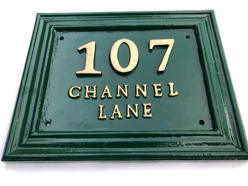 May include: A rectangular, dark green address plaque with gold-colored raised lettering. The plaque reads "107 CHANNEL LANE". The frame has a decorative, layered design. The plaque is likely made of metal or a similar durable material.