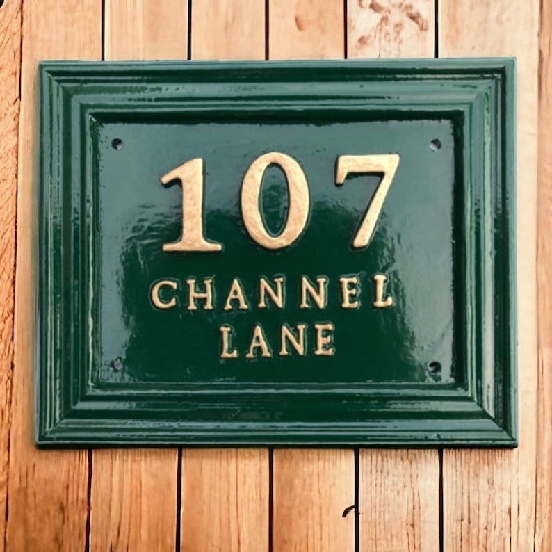 May include: A rectangular, dark green address plaque with the number "107" and the words "CHANNEL LANE" in gold lettering. The plaque has a decorative border and is mounted on a wooden surface.