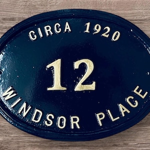 Puede incluir: Una placa de número de casa ovalada azul con letras doradas. La placa dice "Circa 1920" y "Windsor Place" con el número "12" en el centro.
