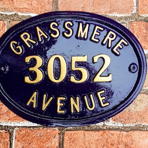 May include: A blue oval-shaped house number sign with gold lettering. The sign reads "Grassmere 3052 Avenue".