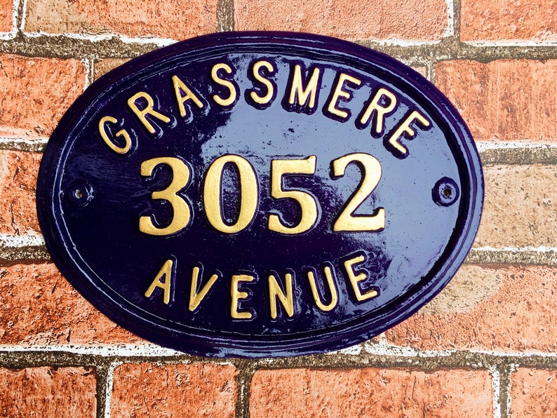 May include: A blue oval-shaped house number sign with gold lettering. The sign reads "Grassmere 3052 Avenue".
