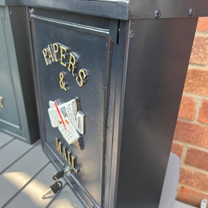 May include: Black metal mailbox with a gold inscription that reads "Papers & Mail". The mailbox has a keyhole and a small, decorative image of a rolled-up newspaper.