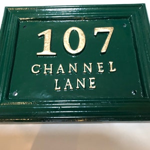 May include: Green rectangular address plaque with the number "107" and the words "CHANNEL LANE" in gold lettering. The plaque has a decorative border and is mounted on a white surface.