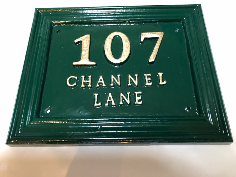 May include: Green rectangular address plaque with the number "107" and the words "CHANNEL LANE" in gold lettering. The plaque has a decorative border and is mounted on a white surface.