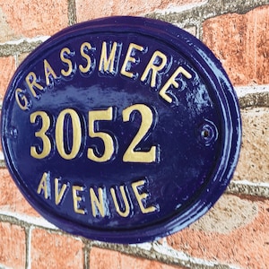 May include: Oval-shaped house number sign with a blue background and gold lettering. The sign reads "GRASSMERE 3052 AVENUE".