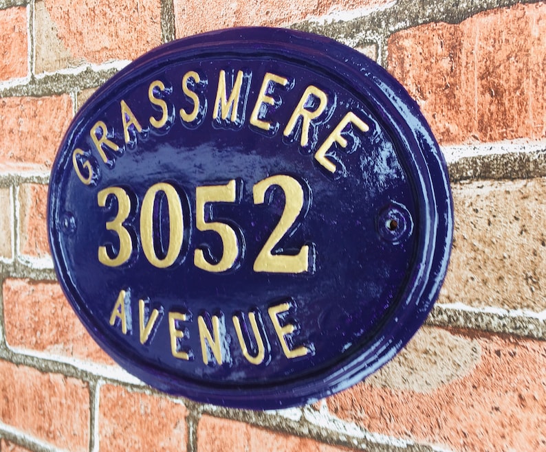 May include: Oval-shaped house number sign with a blue background and gold lettering. The sign reads "GRASSMERE 3052 AVENUE".