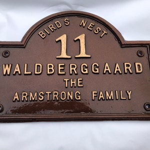Peut inclure: Une plaque en métal brun avec des lettres dorées. La plaque indique "Birds Nest 11 Waldberggaard The Armstrong Family".