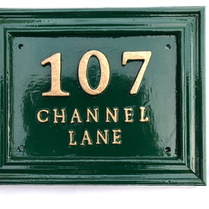 May include: A rectangular, dark green house number sign with gold-colored raised lettering. The sign reads "107 CHANNEL LANE". The sign has a glossy finish and a decorative border. Four screw holes are visible.