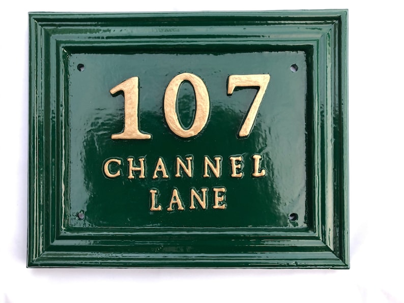 May include: A rectangular, dark green house number sign with gold-colored raised lettering. The sign reads "107 CHANNEL LANE". The sign has a glossy finish and a decorative border. Four screw holes are visible.