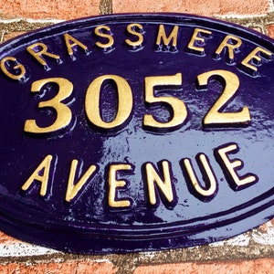 May include: A purple oval-shaped house number sign with gold lettering. The sign reads "Grassmere 3052 Avenue".
