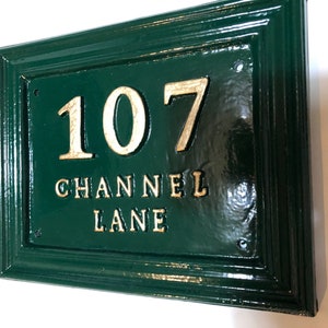 May include: A rectangular, dark green house number sign with the number "107" and the words "CHANNEL LANE" in gold lettering. The sign has a glossy finish and a decorative border. The sign is mounted on a white wall.