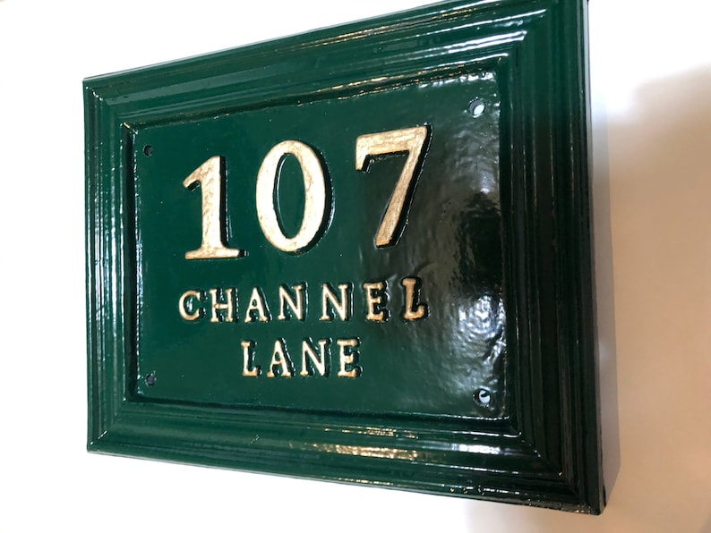 May include: A rectangular, dark green house number sign with the number "107" and the words "CHANNEL LANE" in gold lettering. The sign has a glossy finish and a decorative border. The sign is mounted on a white wall.