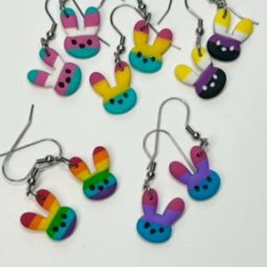 May include: Five pairs of dangle earrings featuring small bunny-shaped charms in various color combinations including rainbow, pink, white, teal, yellow, purple, and black. Each charm has three small dots for eyes and a nose, attached to silver-toned hooks.