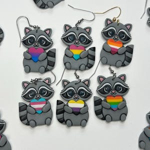 May include: Six pairs of gray raccoon earrings, each featuring a raccoon holding a heart with different colored stripes. The raccoons have black eye masks, white snouts, and striped tails. Each pair hangs from a silver or gold-toned metal hook.