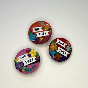 May include: Three circular badges with floral decorations and the text "SHE THEY" on a white ribbon. The badges are in shades of red, purple, pink, yellow, and blue. Each badge is adorned with flowers and leaves, creating a vibrant design.