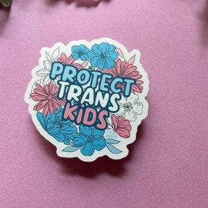 May include: A white sticker with a pink, blue, and white floral design. The sticker says "Protect Trans Kids" in a colorful, stylized font.