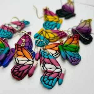 May include: A collection of butterfly-shaped earrings crafted from polymer clay, featuring various vibrant colour gradients including rainbow, pink, white, orange, and blue patterns, each suspended from gold-toned metal earring hooks.