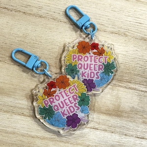 Op de afbeelding: Twee heldere acryl sleutelhangers met blauwe sluitingen. Elke sleutelhanger heeft een bloemenontwerp in regenboogkleuren en de tekst "PROTECT QUEER KIDS". De sleutelhangers liggen op een licht houten oppervlak.