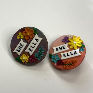 May include: Two round, decorative pins with floral designs and the text "SHE ELLA". One pin is dark purple with orange, yellow, and green flowers. The other is peach with yellow, blue, and orange flowers.