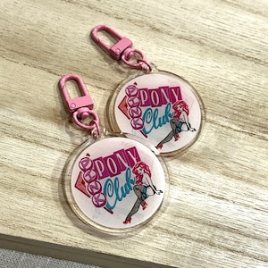 Op de afbeelding: Twee heldere, ronde sleutelhangers met roze hardware. Elke sleutelhanger toont de tekst "PONY Club" in roze en teal, samen met een pin-up illustratie van een vrouw. De sleutelhangers liggen op een licht houten oppervlak.