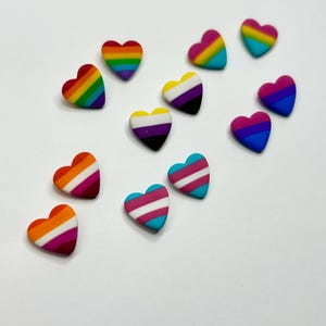 May include: A collection of small heart-shaped charms featuring various horizontal striped patterns in vibrant colors including red, orange, yellow, green, blue, purple, pink, white, and black. Each heart is smooth and displays distinct color combinations.