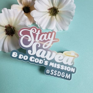 Peut inclure: Un autocollant blanc avec le texte "Stay Saved & Do God's Mission #SSDG" sur un fond dégradé coloré. L'autocollant a une auréole au-dessus du mot "Saved".