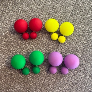 May include: Four pairs of double-sided stud earrings in red, yellow, green, and purple. Each earring has a large, matte sphere with a smaller sphere. The earrings are displayed on a textured, grey surface.