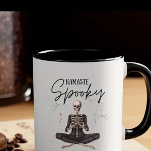 May include: White ceramic mug with a black interior and handle. The mug features the text "NAMASTE Spooky" in a script font, along with an illustration of a skeleton in a yoga pose. The mug is on a wooden surface.