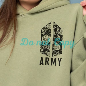 Op de afbeelding: Een lichtgroen hoodie met een zwart bloemenontwerp en de tekst "ARMY" gedrukt op de voorkant.