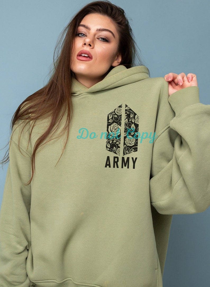 Op de afbeelding: Een lichtgroen hoodie met een zwart bloemenontwerp en de tekst "ARMY" gedrukt op de voorkant.
