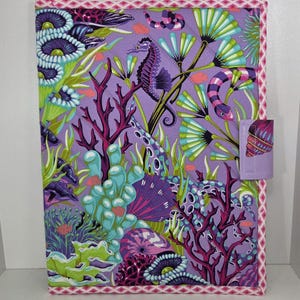 May include: A rectangular quilted fabric cover featuring a vibrant underwater scene on a purple background. The print displays seahorses, coral, sea anemones, and shells in shades of green, pink, and blue. It has a pink and white geometric border.