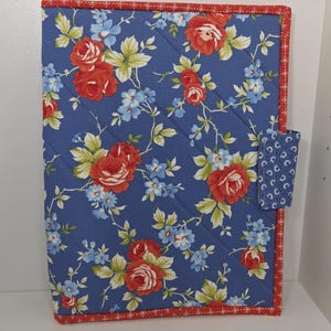 May include: Quilted fabric notebook cover with a floral pattern. The cover features a blue background with red roses and small blue flowers. The edges are trimmed with red fabric.