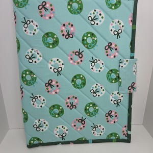 May include: A rectangular quilted fabric cover featuring a light blue background with a repeating pattern of green and white wreaths decorated with pink and black dots and bows. The cover has a dark green fabric border and a small side tab closure.
