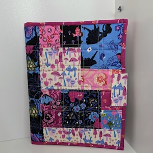 Peut inclure: Une couverture de carnet matelassée avec un motif patchwork. La couverture présente divers carrés de tissu avec des motifs floraux, de champignons et de silhouettes dans des tons de rose, de bleu et de noir. Les bords sont bordés d'un tissu magenta uni.