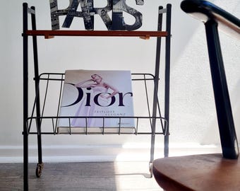 Puede incluir: Un carrito rodante de metal negro con dos estantes. El estante superior tiene un letrero decorativo "PARIS". El estante inferior muestra una revista con la palabra "Dior" en la portada. Una silla de madera está en primer plano.