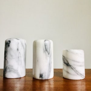 May include: Three white marble cylindrical containers with gray veining. The containers are arranged in a row from largest to smallest on a wooden surface.