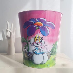 Könnte beinhalten: Rosa Metall-Mülleimer mit einer Cartoon-Illustration einer Maus, die unter einer großen lila Blume schreibt. Die Maus hat eine gelbe Schleife und sitzt auf einem Baumstamm. Der Hintergrund ist ein rosa Himmel und grünes Gras.