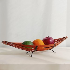 May include: A wooden fruit bowl with a boat-like design, holding a green lime, an orange, and a red apple. The bowl is made of thin, reddish-brown wooden slats and has black metal legs.