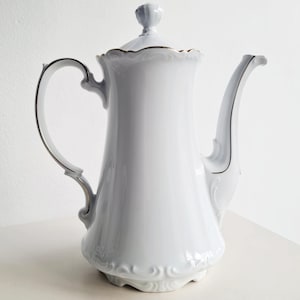 May include: White porcelain coffee pot with a gold trim and a delicate floral design. The pot has a rounded body and a spout on one side.