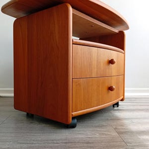 Puede incluir: Una mesita de noche de madera de color marrón claro con una superficie superior curva, un estante de almacenamiento abierto y dos cajones con pequeños pomos redondos de madera. La unidad se apoya sobre cuatro ruedas negras giratorias.