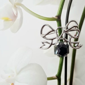 May include: A silver-toned bow brooch with a dark, faceted gemstone center. The brooch is set against a backdrop of white orchid flowers and green stems, creating a delicate and elegant aesthetic.