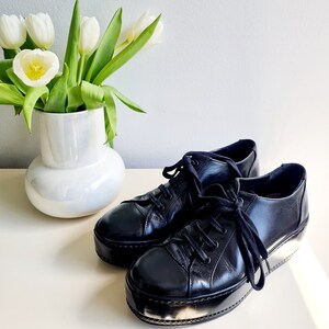 Puede incluir: Un par de zapatillas de plataforma de cuero negro con cordones blancos. Las zapatillas est&aacute;n sobre una superficie blanca junto a un jarr&oacute;n blanco con tulipanes blancos.