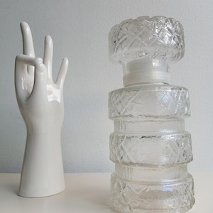 Puede incluir: Una botella de vidrio transparente con un diseño texturizado en forma de diamante. La botella tiene forma cilíndrica con una base ancha y una parte superior decorativa. Una escultura de mano de cerámica blanca está a la izquierda de la botella.