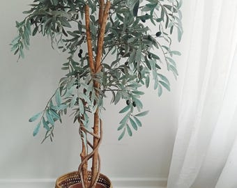 May include: An artificial olive tree with slender, sage-green leaves and small dark fruit clusters. The tree features a braided brown trunk and is housed in a round, woven basket with a geometric pattern in shades of tan, brown, and dark red.