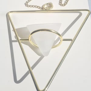 Puede incluir: Un macetero colgante geométrico de metal dorado con un marco triangular. Un jarrón cónico de vidrio esmerilado está suspendido dentro del marco. Una delicada cadena y un anillo están unidos para colgar. El macetero es para plantas pequeñas.