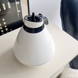 May include: A white ceramic teapot with a black lid and handle. The teapot has a rounded, bulbous shape.