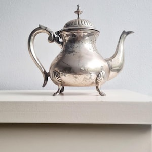May include: A vintage silver teapot with a curved handle and spout. The teapot has a lid with a decorative finial and rests on three small feet. The body of the teapot has a bulbous shape with decorative shell-like accents.