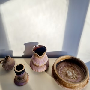 Puede incluir: Un conjunto de cuatro piezas de cerámica en tonos marrones y morados. El conjunto incluye una jarra, un tazón pequeño y dos jarrones pequeños.