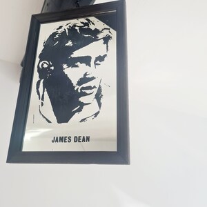 May include: A black and white framed mirror with a silhouette of James Dean's face and the text "JAMES DEAN" printed below the image.